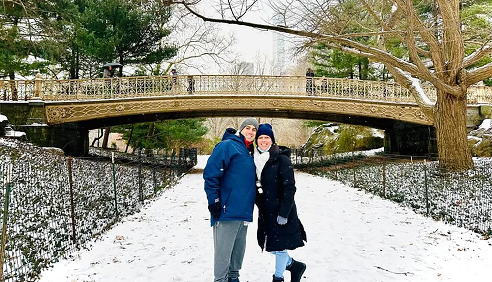 A smiling couple poses for a photo in front of an ornate bridge in a snowy park setting