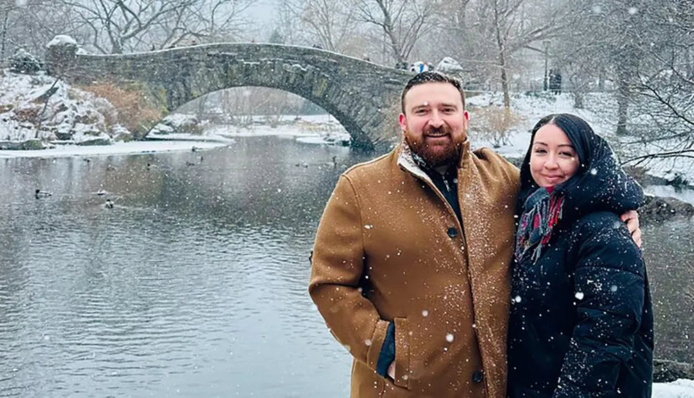 Two people pose for a photo in front of a picturesque stone bridge over a partially frozen river in a snowy landscape