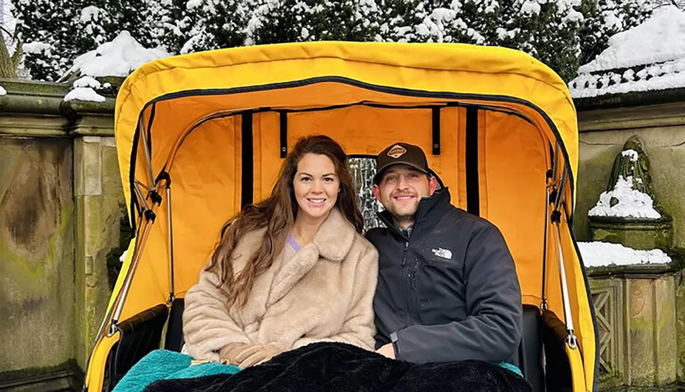 A smiling man and woman are sitting together in a yellow horse-drawn carriage with a snowy backdrop