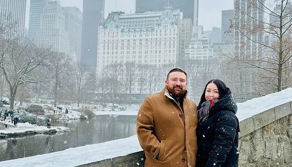 A couple poses for a photo with smiles on a snowy day in an urban park setting with snow-covered trees and buildings in the background