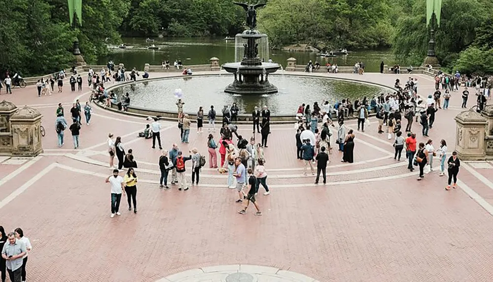 The image shows a bustling public space with a central fountain where many people are gathering and walking around likely in a park setting