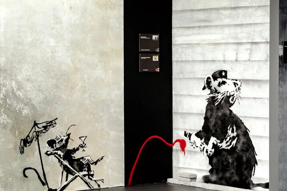 The image displays a wall with two graffiti artworks one depicting a rat with a cap holding a marker and the other showing a person being pushed in a shopping cart by a skeleton with a red line seemingly connecting both graffiti works