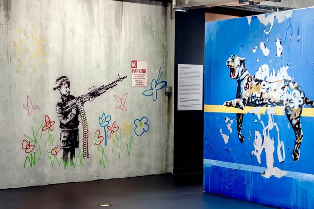 The image shows an exhibition space with two walls featuring street art to the left theres a piece showing a child with a gun that shoots out flowers and to the right a multicolored stencil of a dog is displayed on a peeling blue background