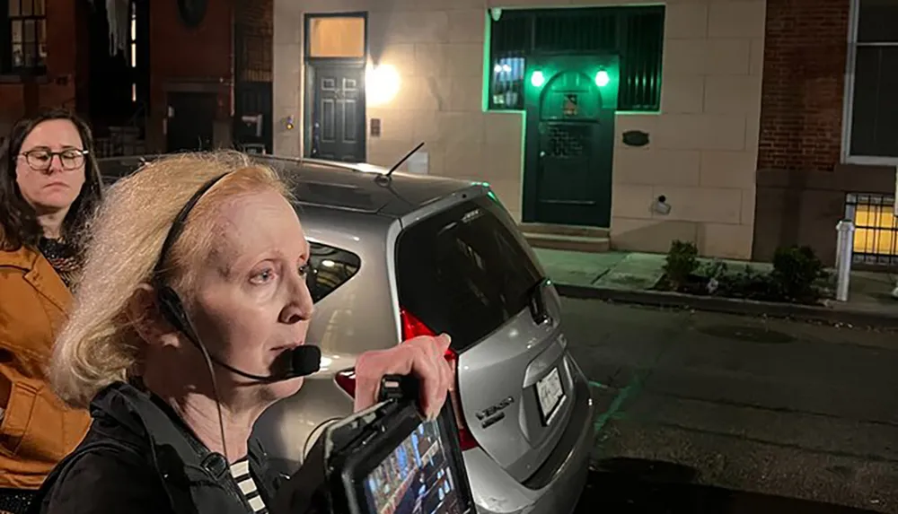 A person wearing a headset is holding a device with multiple screens during a nighttime setting with another individual and a car visible in the background