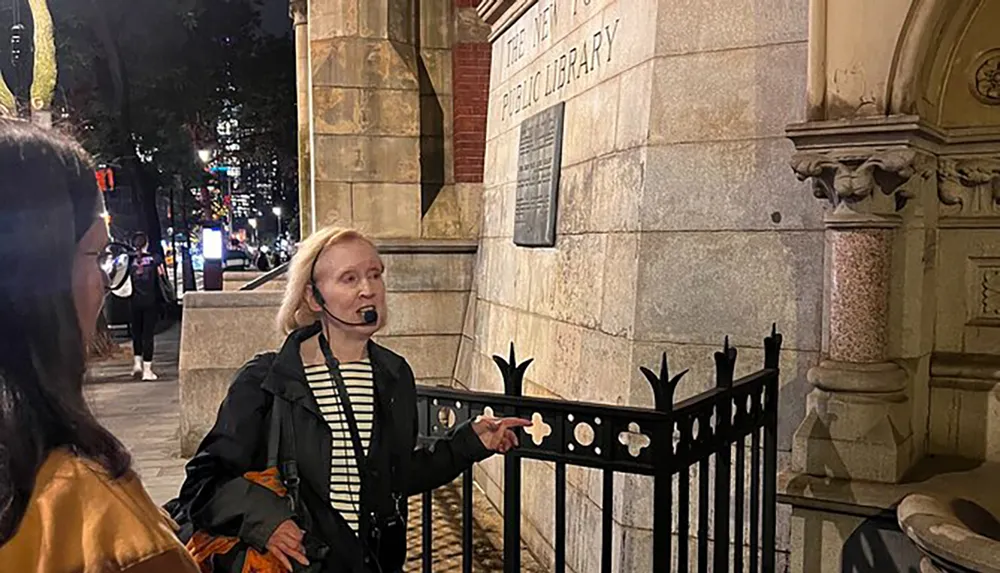 A tour guide is speaking to a visitor outside of a building that appears to be the New York Public Library at night