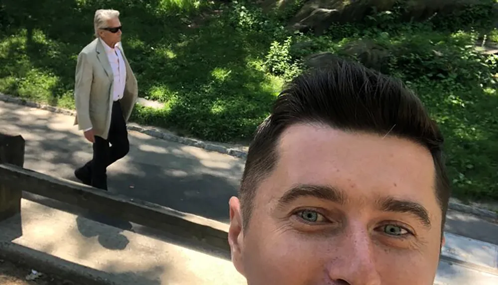 A man is taking a selfie with another person unintentionally captured in the background walking on a path surrounded by greenery