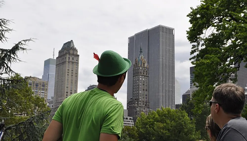 A person wearing a green hat with a feather is overlooking a park with skyscrapers in the background