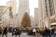 The image shows a bustling crowd of people gathered around a large decorated Christmas tree in an urban setting, indicative of a holiday celebration in a city.