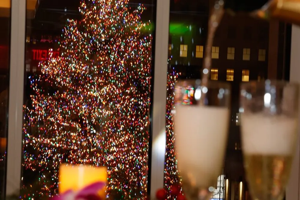 The image shows an intricately lit Christmas tree viewed from an indoor setting with the trees twinkling lights creating a festive atmosphere through a window-pane accompanied by two glasses of what appears to be a bubbly beverage in the foreground