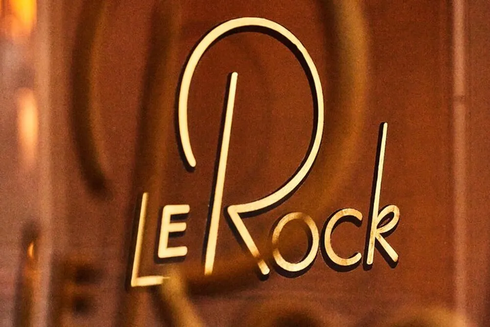 The image shows a close-up of a stylish golden sign that reads LE ROCK against a blurry background