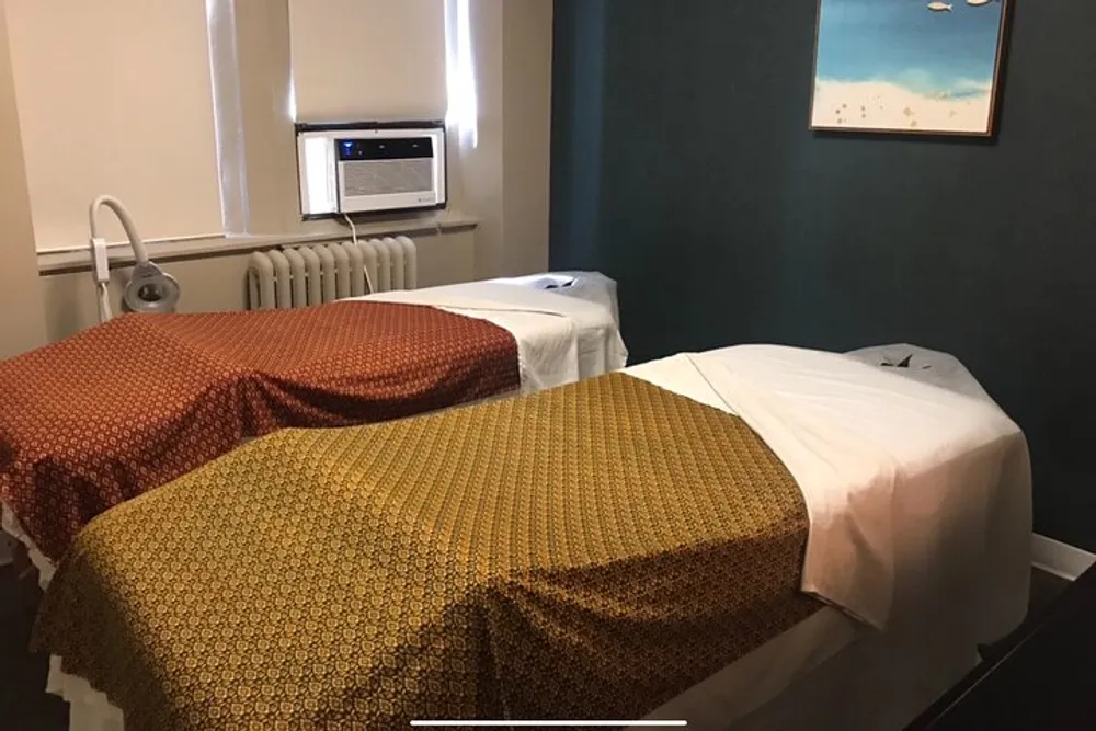 The image shows a room with two massage tables covered with patterned towels a serene wall color and a piece of art hanging on the wall suggesting a spa or wellness center environment