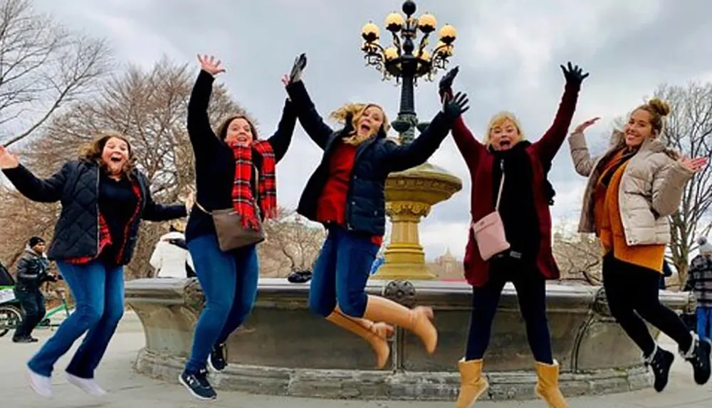 A group of five people joyfully jump into the air with their arms raised in front of a lamp post and fountain capturing a lively moment outdoors