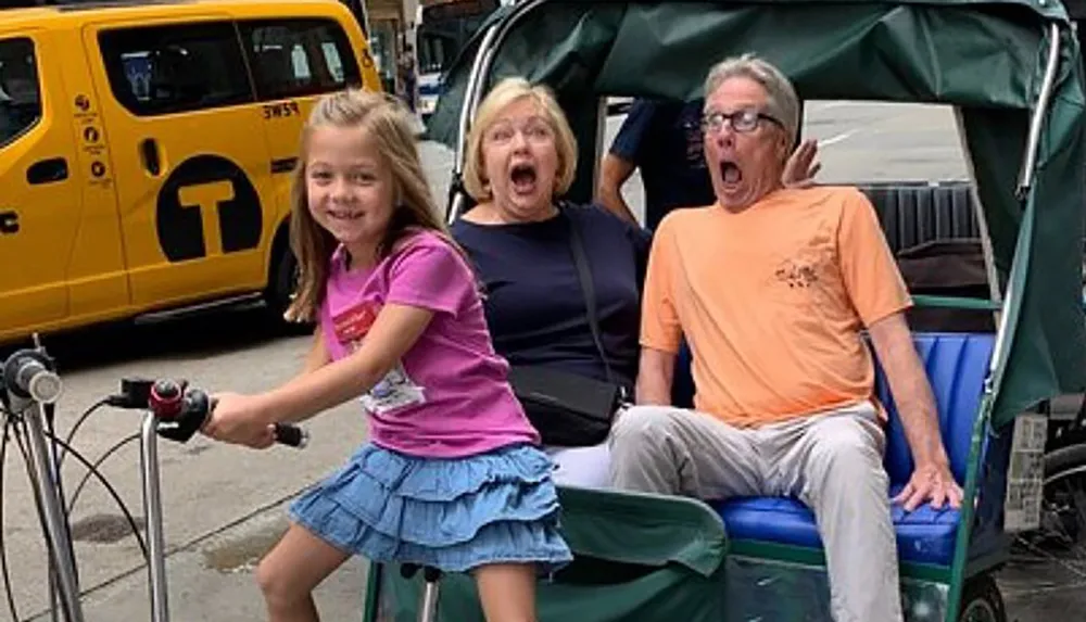 A young girl is playfully pretending to steer a rickshaw bike with two adults showing exaggerated shocked expressions in the back seat