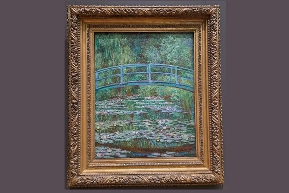 The image displays an impressionist painting featuring a tranquil pond with lily pads and a Japanese-style wooden bridge all surrounded by lush greenery and framed in an ornate gold frame