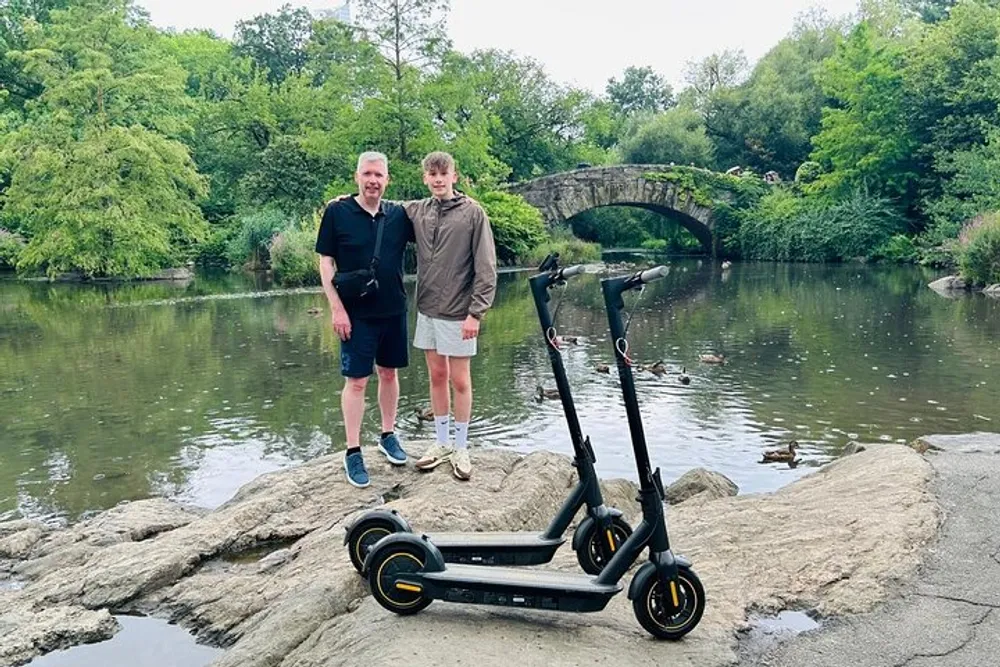 Two individuals and two electric scooters are standing on a rock with a pond and an arched bridge in the background