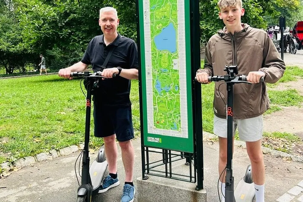 Two people are smiling and standing with electric scooters next to a map sign in a park-like environment