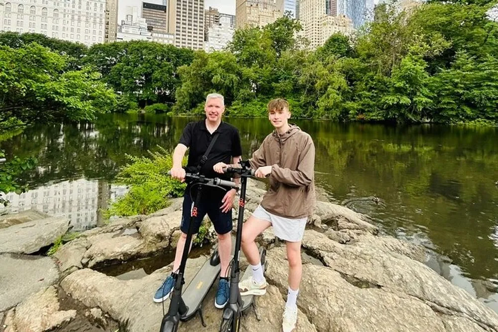 Two individuals are standing on a rock by a pond in an urban park with scooters smiling for the photo