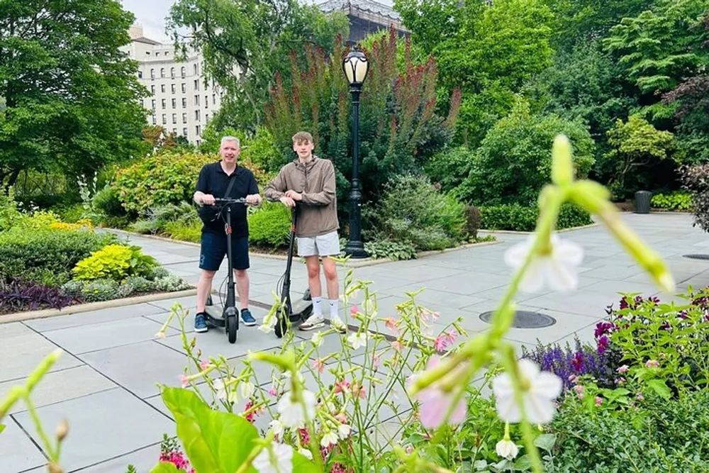 Two individuals are standing with electric scooters in a lush park with blooming flowers in the foreground