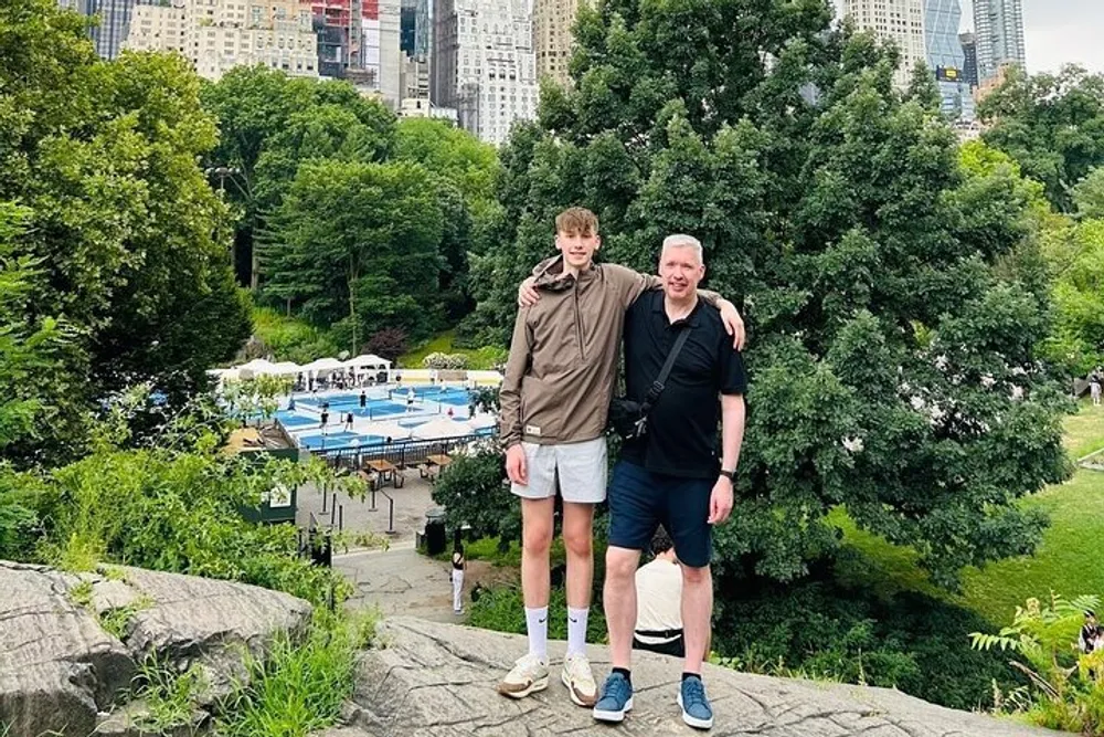 Two people are standing together on a rock overlooking a swimming pool and greenery with city buildings in the background likely taken in a park setting