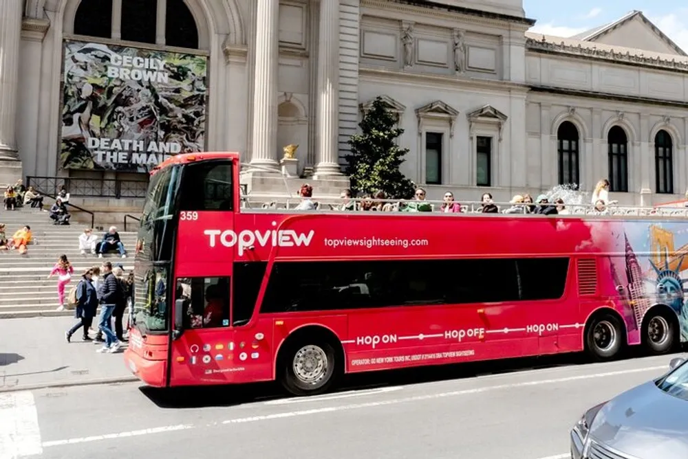 A red double-decker sightseeing bus marked TopView is parked in front of a neoclassical building with people sitting on top and others milling about indicating a bustling city scene likely popular with tourists