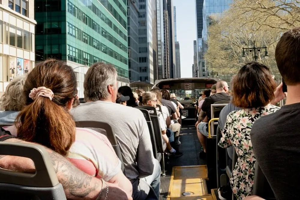 Passengers are seated on an open-top tour bus driving through a city with towering skyscrapers lining the street