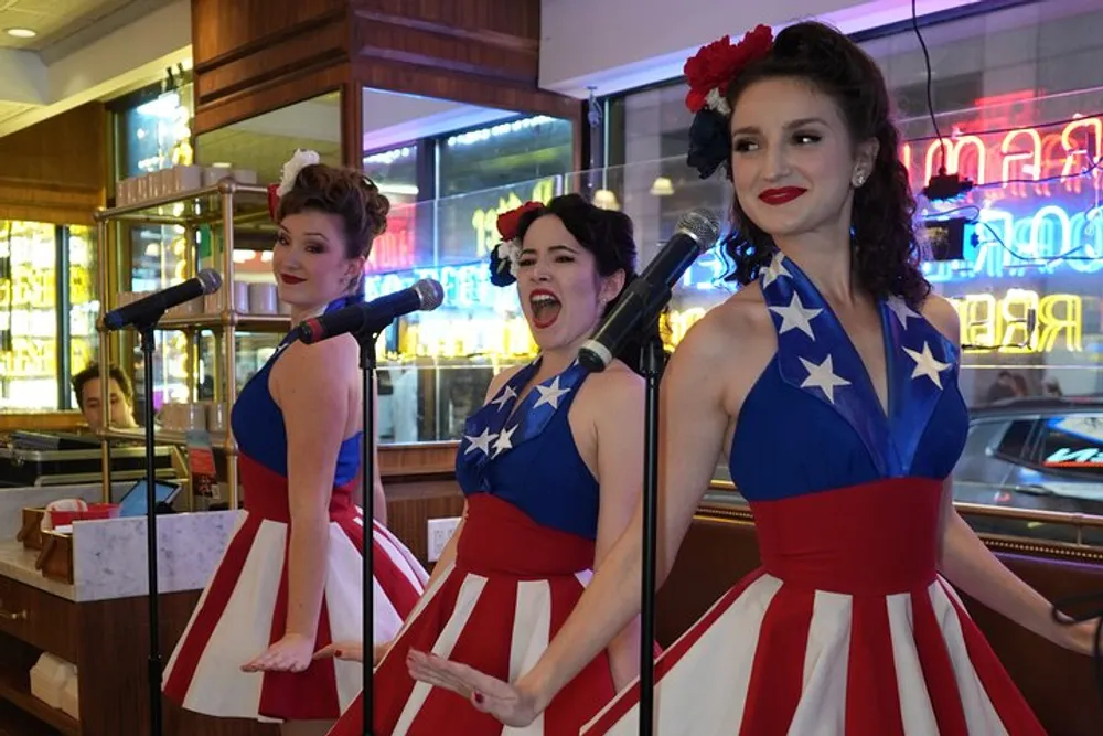 Three women wearing vintage-style dresses with an American flag motif are singing into microphones evoking a classic Americana performance vibe