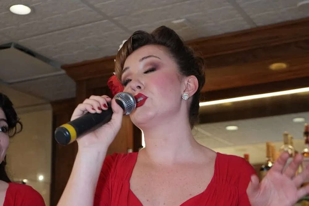 A woman in a red dress with a vintage hairstyle is singing into a microphone eyes closed conveying a strong emotion through her performance