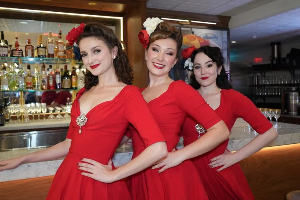 Three women in matching red dresses and flower hair accessories are smiling in front of a bar