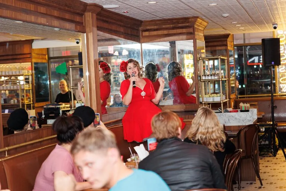 A person in a red dress is performing or speaking into a microphone in front of an audience at an indoor venue