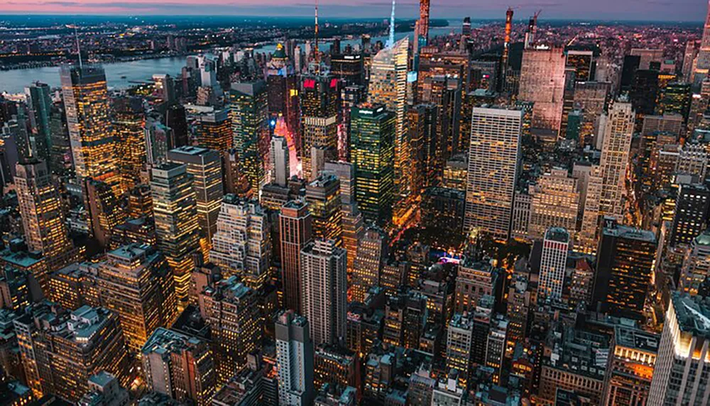 The image captures an aerial view of a densely packed urban skyline at dusk illuminated by the vibrant lights of skyscrapers and streets