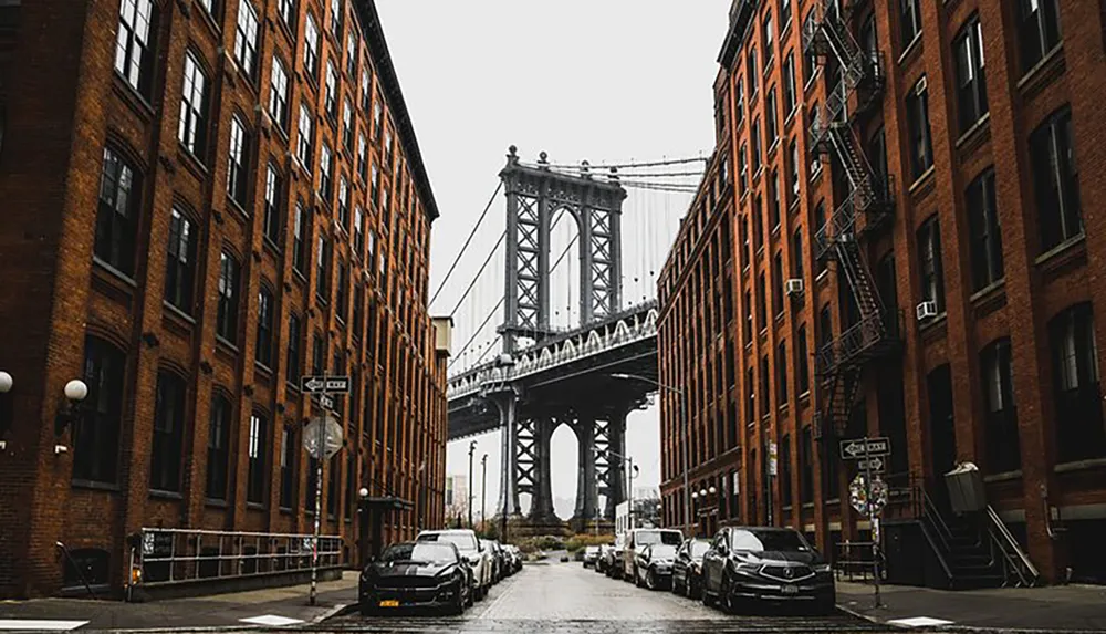 A moody overcast view down a cobblestone street flanked by brick buildings with the Manhattan Bridge looming in the background