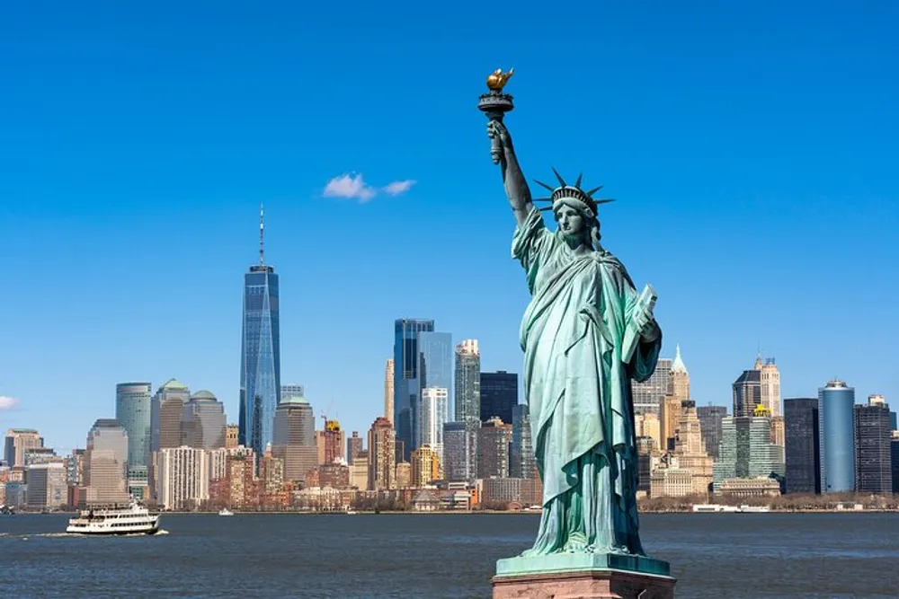 The Statue of Liberty stands prominently in the foreground with the skyline of Lower Manhattan and a clear blue sky in the background