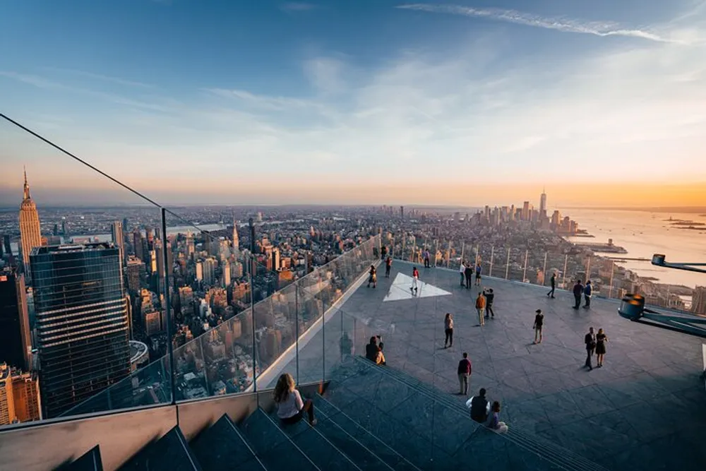 Visitors enjoy a panoramic view of the New York City skyline at sunset from a high vantage point with clear glass barriers