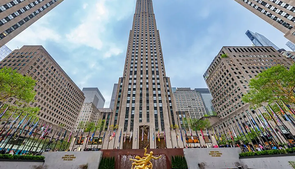 The image shows a view of Rockefeller Center with its towering skyscraper and the iconic golden Prometheus statue in the foreground surrounded by a colorful array of international flags