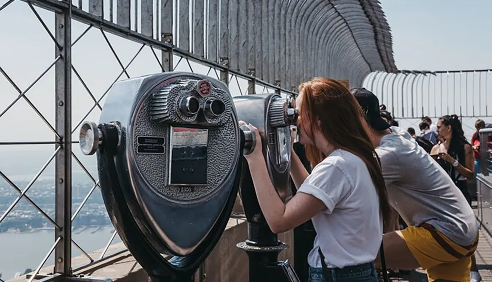 A person is looking through a coin-operated binoculars at a high vantage point with other visitors around