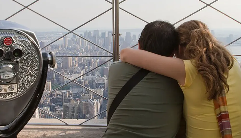 Two people are embracing while looking out over a cityscape from a high vantage point with a coin-operated binocular viewer in the foreground