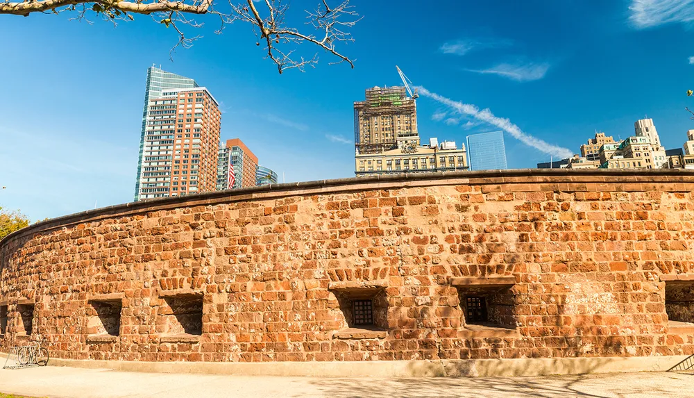 The image shows an old rounded stone fortification with multiple openings contrasted by a backdrop of modern high-rise buildings and a clear blue sky