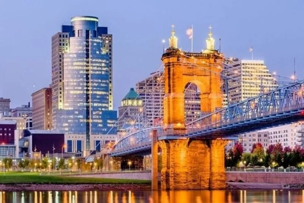 The image captures a lit-up stone bridge with intricate arches spanning a river leading towards a modern city skyline illuminated against the dusk sky