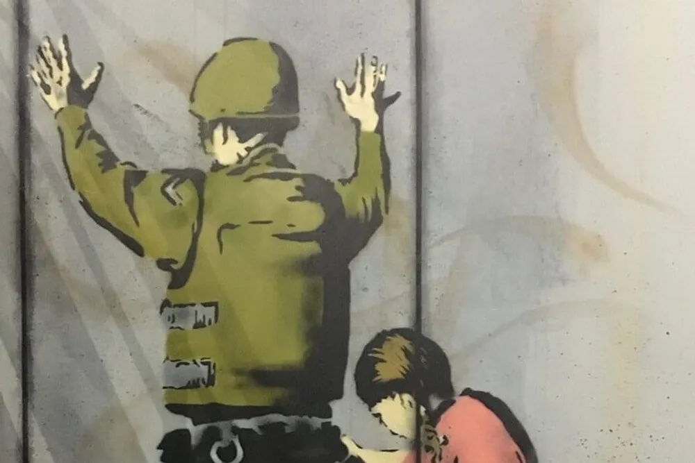 This image depicts a stenciled graffiti artwork showing a person in military attire with their hands up against a wall while another individual crouches behind them