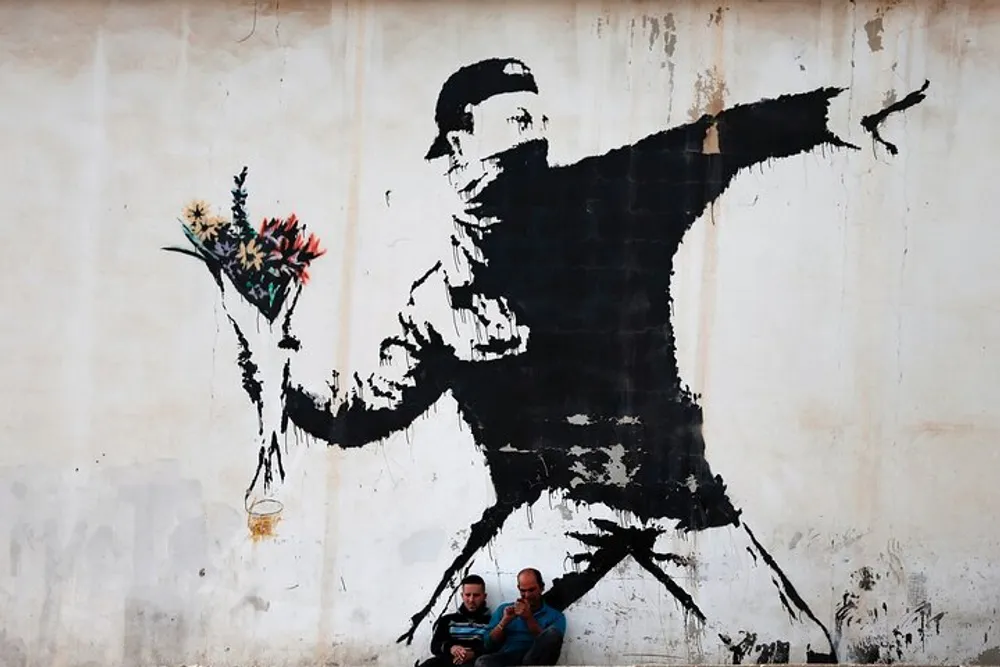 Two individuals are seated beneath a large monochromatic street art mural that depicts a person throwing a bouquet of flowers