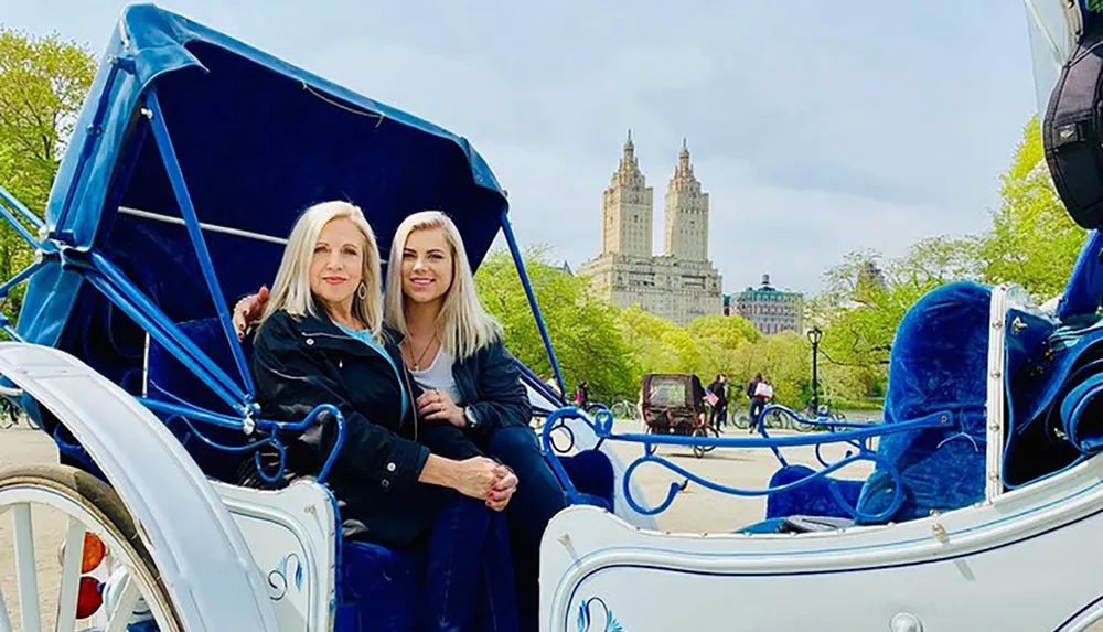 Two women are smiling and posing together in a horse-drawn carriage with a scenic background that includes lush greenery and towering buildings