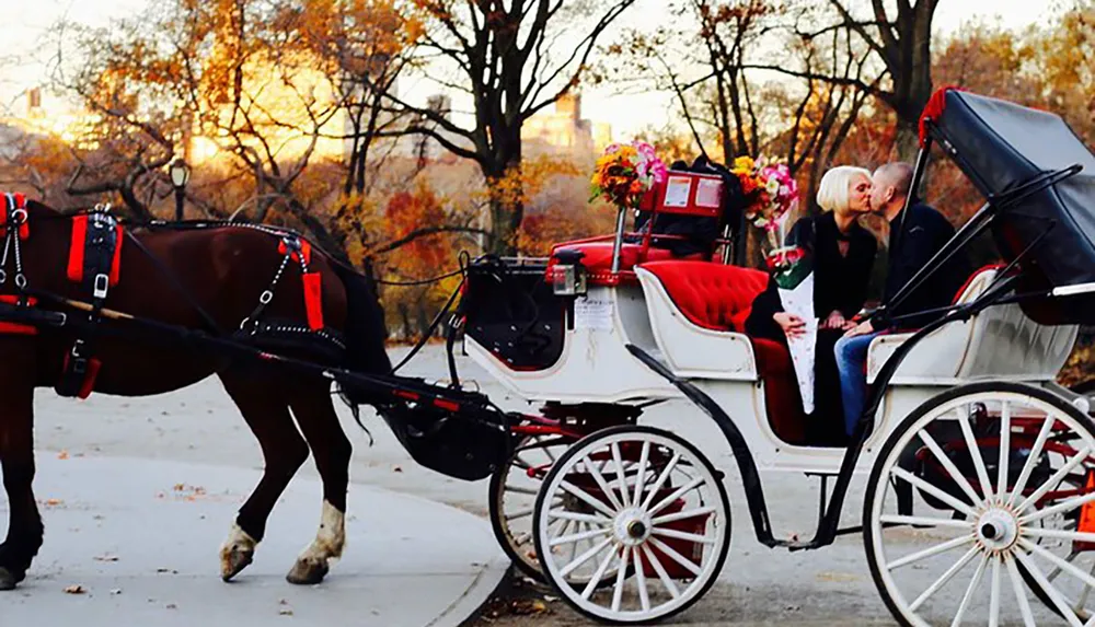 Two people share a kiss in a horse-drawn carriage adorned with flowers set against a backdrop of autumn foliage