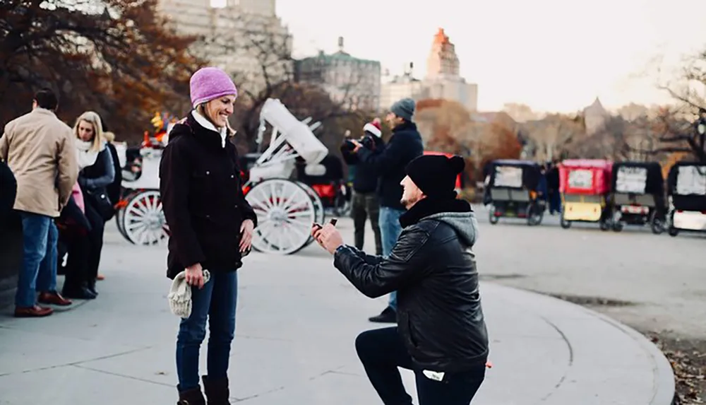 A person is proposing to their partner on a pathway with bystanders in the background creating a moment of joy and surprise