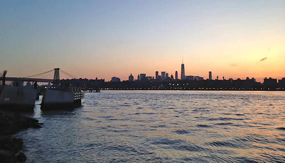 The image captures a tranquil sunset over a river with a silhouette of a city skyline and a bridge in the background