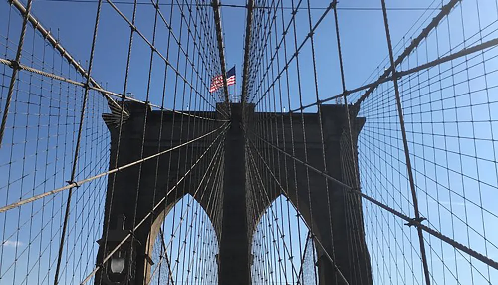 The image shows a close-up view of the intricate cable pattern of the Brooklyn Bridge with an American flag flying at its pinnacle against a clear blue sky