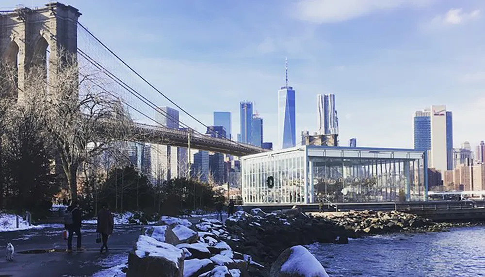 The image captures a winter scene with the Brooklyn Bridge and the Manhattan skyline including the One World Trade Center in the background as people walk by the riverfront likely in Brooklyn Bridge Park with a dog and patches of snow visible on the ground