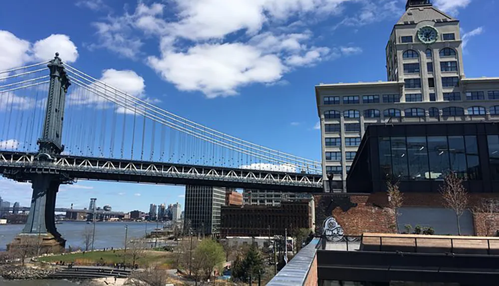 The image features a picturesque view of the Manhattan Bridge spanning over a river with a blue sky above adjacent to an elegant building with a clock tower