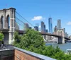 This image captures the iconic Brooklyn Bridge with its distinctive stone towers and cable arrangement in the foreground while the modern skyscrapers of Lower Manhattan including One World Trade Center create a contrasting backdrop under a clear blue sky