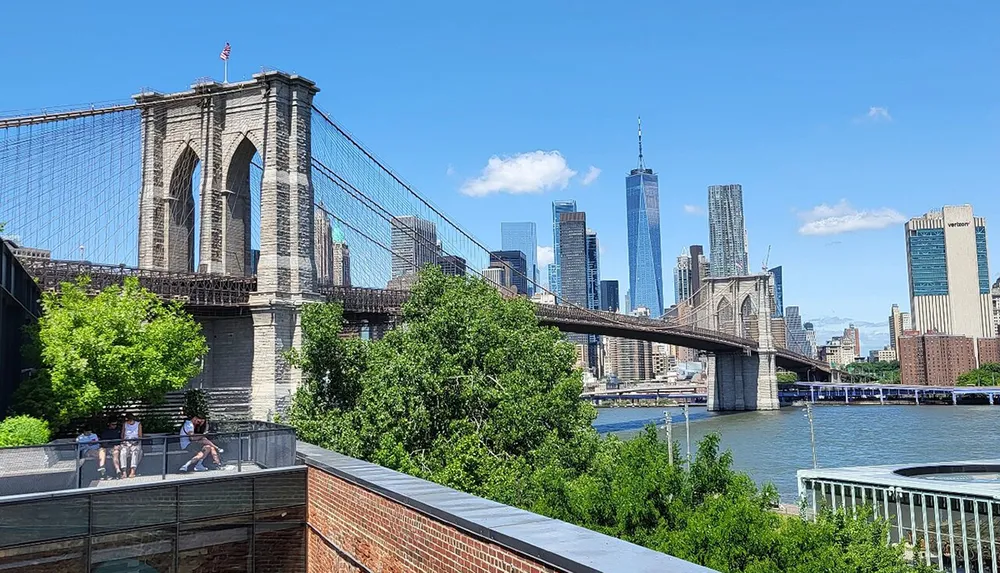 This image captures the iconic Brooklyn Bridge with its distinctive stone towers and cable arrangement in the foreground while the modern skyscrapers of Lower Manhattan including One World Trade Center create a contrasting backdrop under a clear blue sky