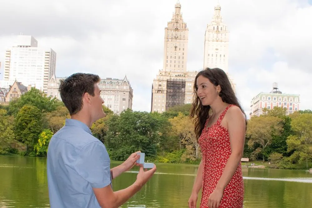 A person is proposing to their partner with a ring by a lake with city buildings in the background